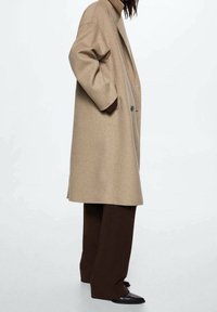 Beige wollen jas met een ontspannen pasvorm, dubbelgeknoopt ontwerp en brede revers, gecombineerd met bruine wijde broekspijpen en puntige zwarte schoenen.