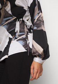 Blouse noire et crème avec un motif floral abstrait, manches longues et un lien à la taille. Tissu lisse avec un léger éclat.