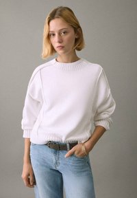 Bela, teksturirana majica z rebrastim izrezom in manšetami, kombinirana s svetlo modrimi jeans hlačami in rjavim pasom. Modelka pozira pred sivim ozadjem.