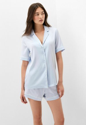 Vrouw draagt lichtblauw kort mouw knoophemd en bijpassende korte broek, staand tegen een effen witte achtergrond.