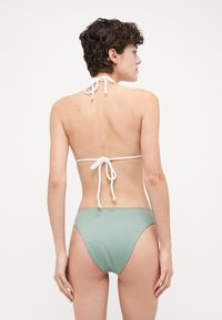 Žalias banguotas bikini, turi kaklo užrišimą su baltomis juostomis, atvirą nugarą ir minimalistinį stilių. Subtilus aukso akcentas gale.