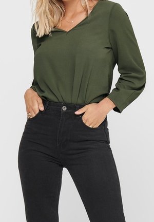 Kobieta w oliwkowozielonej bluzce z długim rękawem i dekoltem w serek, włożonej do czarnych, wysokowaistowych dżinsów, z rękami w kieszeniach.