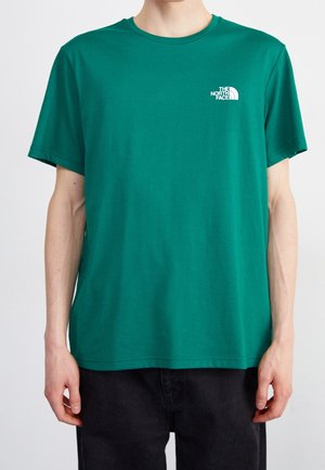 Person trägt ein einfarbig grünes T-Shirt mit kurzen Ärmeln und The North Face-Logo auf der linken Brust, kombiniert mit schwarzen Hosen.
