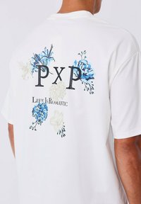 Camiseta blanca de manga corta con gráficos florales en azul y crema, con "PXP" y "La vida es romántica" impresos en negro.