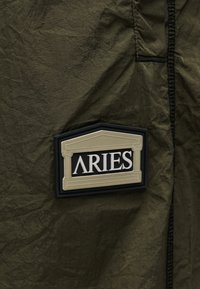 Μπαλώματα με κείμενο «ARIES» σε λευκό χρώμα σε μαύρο και μπεζ φόντο, ραμμένα σε τσαλακωμένο λαδί ύφασμα με ορατή μαύρη ραφή.