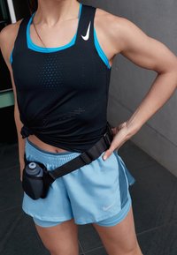 Canotta nera Nike con accenti blu, dotata di perforazioni. Abbinata a pantaloni corte blu chiaro e una borsa nera in vita che tiene una bottiglia d'acqua.