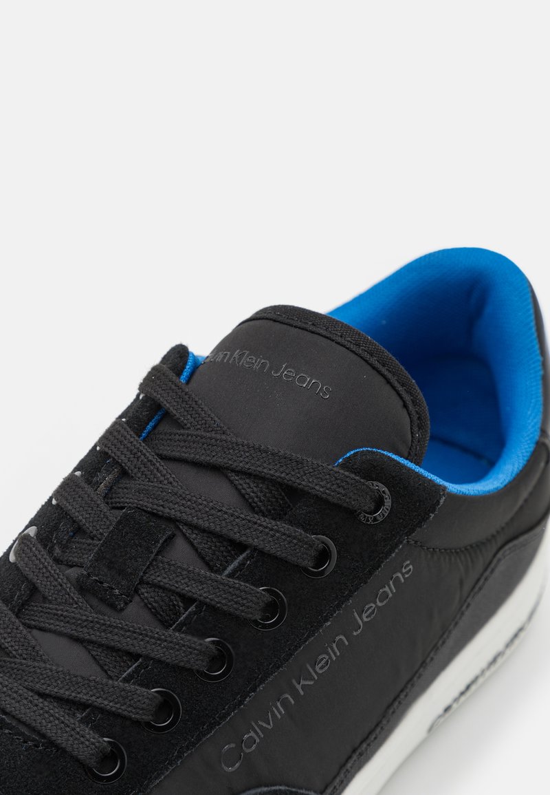 Chaussure de sport noire avec un dessus en daim et textile. Caractéristiques d'un doublure bleue, de lacets noirs et d'une marque en relief sur la languette. Semelle blanche.