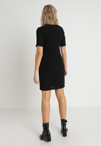 Svart klänning med korta ärmar, figursydd design och knälång längd. Har dragkedja baktill och slät textur. Stylad med glänsande svarta stövlar.