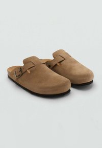 Sabots en suede marron avec un bout rond, une sangle à boucle ajustable et une semelle en caoutchouc noir texturé, offrant un design minimaliste.