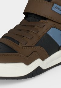 Gros plan d'une basket marron, noire et bleue avec des lacets, une sangle en velcro et une semelle blanche avec une bande de roulement noire.