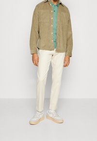 Muž v béžové kordové bundě, tyrkysovém polo triku, bílých kalhotách a bílých teniskách, stojí proti jednoduchému světlému pozadí.