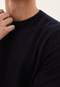 Marineblauer Strickpullover mit einem gerippten Rundhalsausschnitt und weicher Textur. Nahaufnahme des Kragens und des Teilarms, die die Nähdetails hervorhebt.