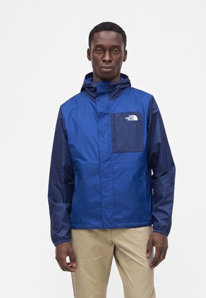 Mand iført en blå North Face jakke med hætte og beige bukser, stående mod en ensfarvet lys baggrund.