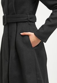 Schwarzer Wollmantel mit einem taillierten Design, seitlichen Taschen und einem Gürtel in der Taille. Strukturiertes Material mit glatter Oberfläche. Lange Ärmel.