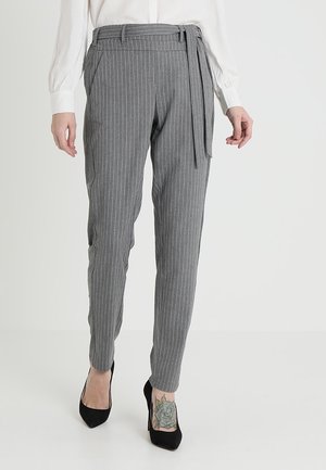 Pantalon gris à fines rayures fuselé avec ceinture à la taille, porté avec des talons noirs et un chemisier blanc à manches longues sur un fond uni.