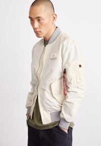 Giacca bomber di crema chiara con polsini e collare a coste grigi. Presenta una tasca laterale con zipper rossa e un patch sul petto sinistro.