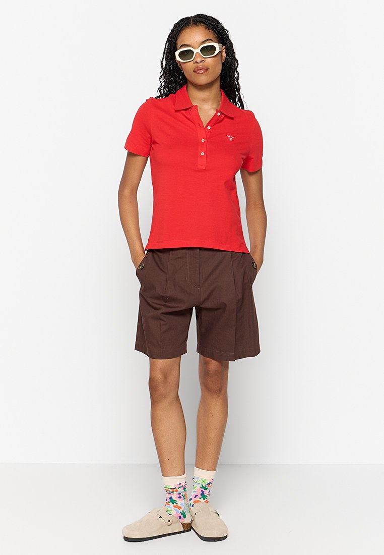 Camisa polo roja de mangas cortas con un frontal abotonado, combinada con pantalones cortos marrones. Los accesorios incluyen gafas de sol blancas y calcetines con un diseño colorido.