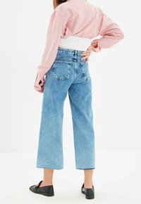 Nainen selin punaisessa pitkähihaisessa paidassa, leveälahkeisissa sinisiä farkuissa, joissa on raakapäät, mustissa loafer-kengissä ja valkoisessa selkätuesta.