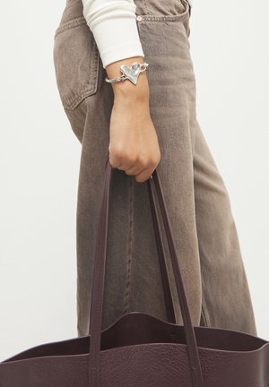 Persona con pantalones beige y manga blanca sosteniendo un bolso tote de cuero marrón oscuro, con una pulsera plateada en forma de corazón en la muñeca.