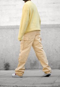 Stickad gul tröja och creamfärgade cargobyxor. Byxorna har sidfickor; skorna är vita med röda detaljer. Bakgrunden är en suddig betongyta.