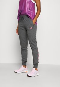 Grijze joggingsbroek met een tapere pasvorm, roze Adidas-logo op de dij, gecombineerd met een paarse mouwloze top en roze sportschoenen.