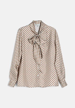 Vrouw met steil donker haar draagt een blouse met patroon en strikceintuurkraag en een bruine broek, staat met handen achter haar rug tegen een witte achtergrond.