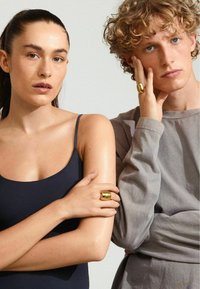 Anillos de oro en las manos; la mujer lleva una camiseta sin mangas azul marino y el hombre tiene una camisa de manga larga gris claro, ambos contra un fondo neutro.