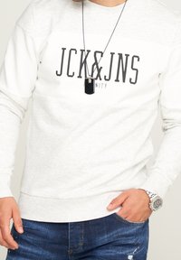 Světle šedý svetr s tmavě šedým nápisem "JCK&JNS" vpředu, kulatý výstřih a žebrované manžety. Spárováno s tmavě modrými džíny a hodinkami.