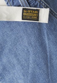 Kék farmeranyag textúrázott felülettel; fehér és fekete G-Star Originals Raw Denim címke varrva a derékpántra.