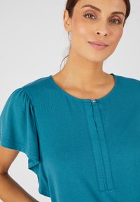 Top azul verdoso con mangas cortas abullonadas, escote redondo, detalle de botones en el frente y una textura suave y lisa. Diseño casual y versátil.