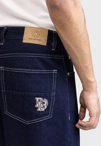 Nahaufnahme von dunkelblauen Denim-Jeans mit weißen Nähten, einem braunen "Redefined Rebel"-Label am Bund und der gestickten Aufschrift "RR Legendary" auf der Gesäßtasche.