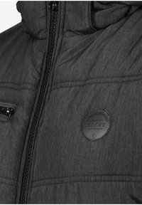 Polstret grå jakke med teksturert overflate, med glidelås foran, sidelomme og et rundt lærlogo-patch på brystet.