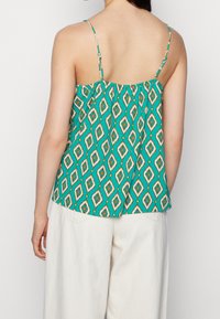 Débardeur vert à motif avec un design géométrique, fines bretelles réglables et coupe ample. Porté avec un pantalon large de couleur claire.