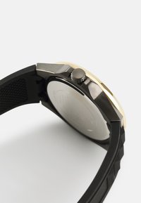Bracelet de montre en caoutchouc noir avec surface texturée, boîtier en métal doré et couronne latérale. Le dos de la montre présente une surface lisse et réfléchissante.