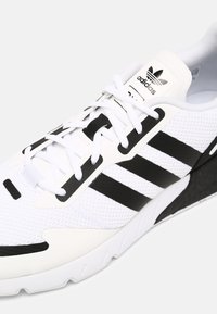 Adidas apavi baltā acs audumā ar melnām svītrām un akcentiem. Noapaļota purngalu forma, mīksts apkakle un teksturēta zole. Šņorējumi redzami priekšpusē.