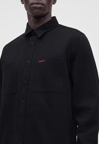 Homme portant une chemise noire à boutons avec deux poches poitrine, dont une avec un petit texte rouge "HUGO" sur un fond uni gris clair.