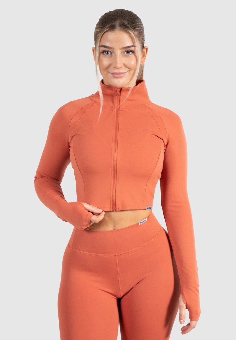 Gekürzte orangefarbene Zip-Jacke aus dehnbarem Material mit langen Ärmeln, die Daumenlöcher und eine figurbetonte Passform aufweist. Kombiniert mit hochgeschnittenen Leggings.