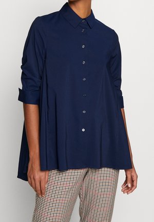 Persona con una camisa azul oscuro de botones y suelta, con las mangas remangadas, y pantalones a cuadros, frente a un fondo claro y liso.