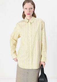 Camicia a righe gialle con linee viola, maniche lunghe e colletto. Abbinata a una gonna di jeans chiara e una borsa nera.