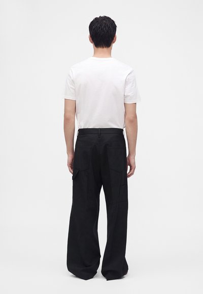 Homme de dos portant un t-shirt blanc uni à manches courtes et un pantalon noir ample avec des poches arrière sur un fond clair uni.