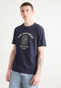 Mørkeblå bomullst-skjorte med rund hals, med frontgrafikk med teksten "GANT SPORTSWEAR" og "NEW HAVEN 1949" i beige.
