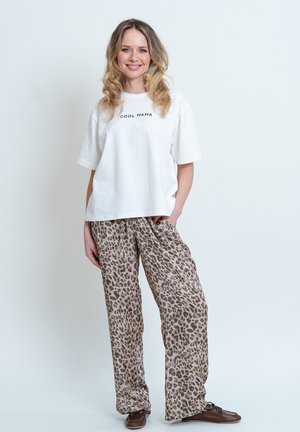 T-shirt bianco in cotone con stampa "COOL MAMA", abbinato a pantaloni beige a vestibilità larga con design a stampa leopardata e scarpe marroni.