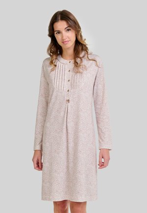 Mujer con cabello castaño ondulado que lleva un vestido rosa claro de manga larga y hasta la rodilla, con un pequeño patrón de puntos y botones en el pecho.