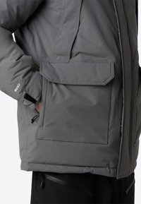 Grå isolerad jacka med en texturerad yta, stor framficka och justerbara ärmslut. Inkluderar en dragsko i nederkanten.