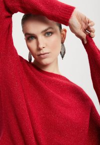 Roter Strickpullover mit lässiger Passform, glitzernder Textur und langen Ärmeln. Das Model trägt hängende silberne Ohrringe und hat zurückgegeltes Haar.