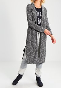 Cárdigan largo y abierto en una textura gris jaspeada, combinado con una camiseta gráfica negra y pantalones gris claro, complementados por botas de tobillo.