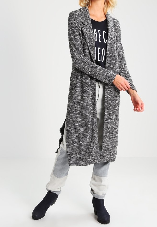 Cárdigan largo y abierto en una textura gris jaspeada, combinado con una camiseta gráfica negra y pantalones gris claro, complementados por botas de tobillo.