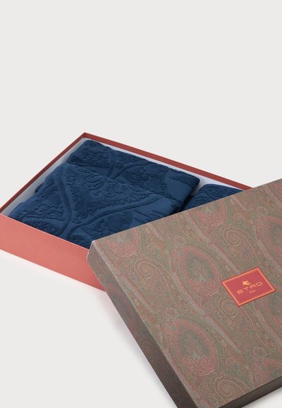 Deux serviettes bleu foncé pliées avec des motifs en relief à l'intérieur d'une boîte cadeau partiellement ouverte à motifs avec une étiquette Etro Home.