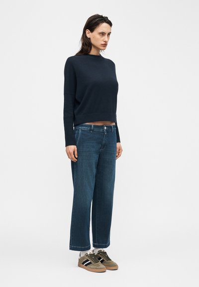 Femme portant un pull bleu marine, un jean large bleu, des chaussettes blanches et des baskets vert olive, debout devant un fond blanc.