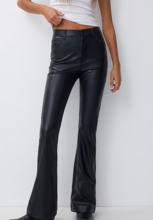 Woman wearing high-waisted black faux leather flare pants and a white cropped tank top, standing against a white background.